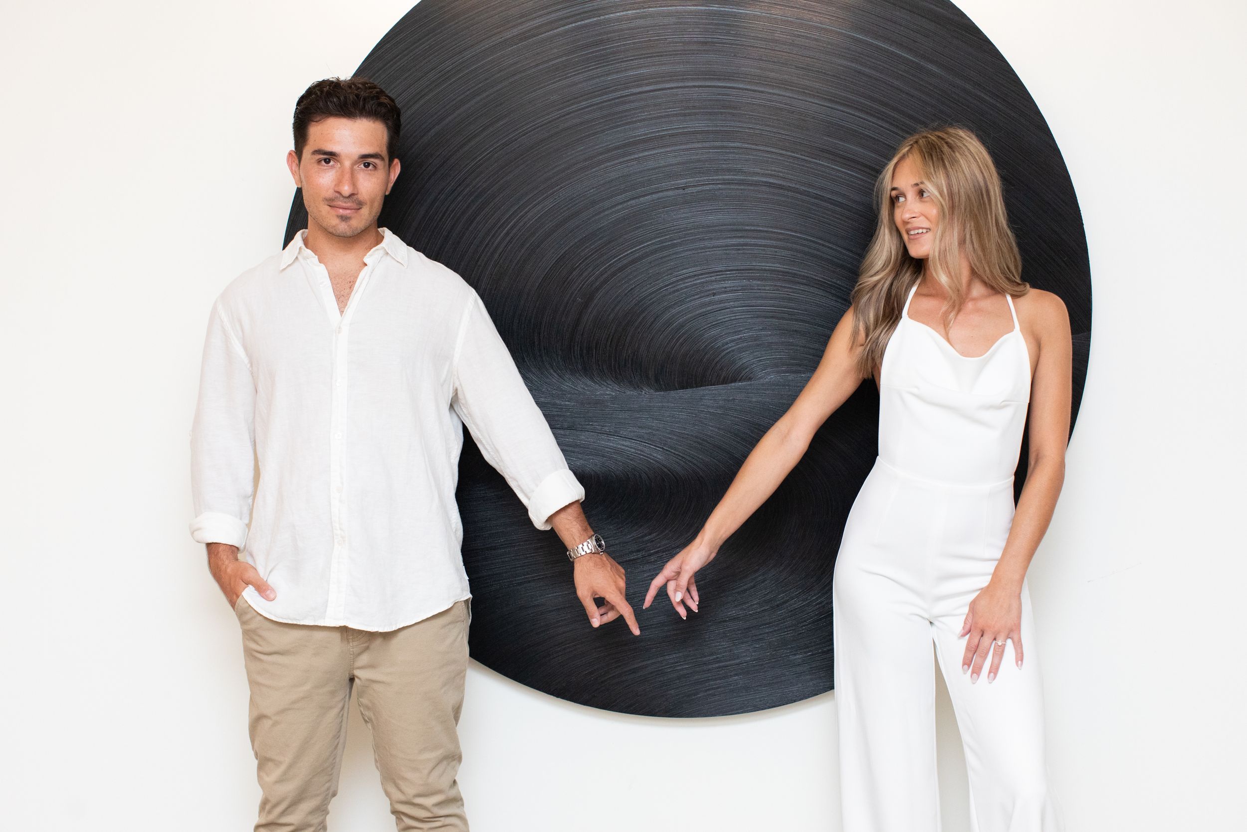 A stylish couple in white holds hands against a black circular wall backdrop.