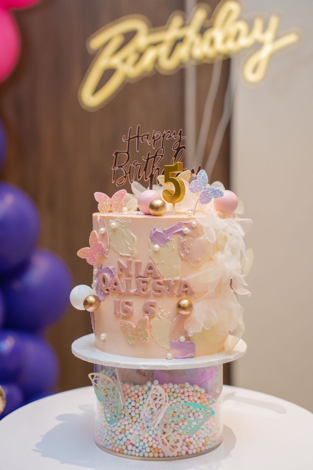 Pink and white birthday cake with gold decor and neon birthday sign on display.