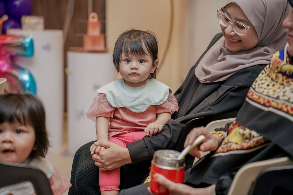 Young child in pink and white outfit sitting on lap at indoor celebration.