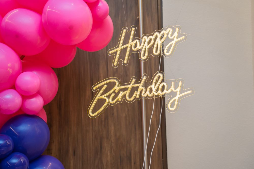 Neon Happy Birthday sign with pink and purple balloon decoration.