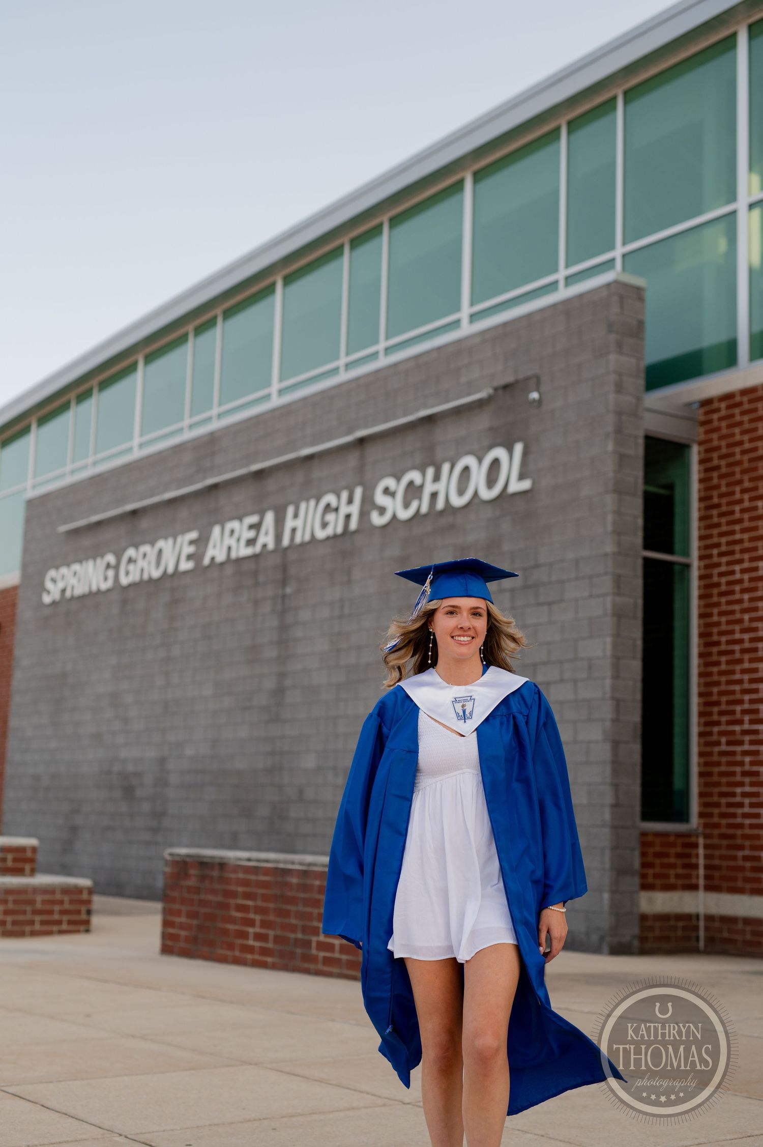 Graduate in blue regalia poses in front of high school building signage.