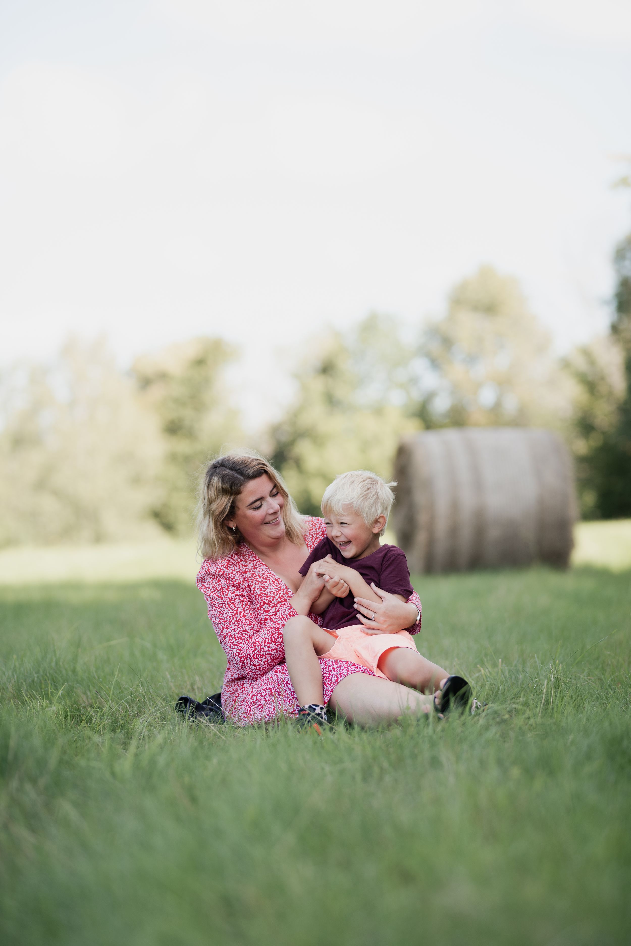 Family members play and laugh together on grassy field with hay bale in background.