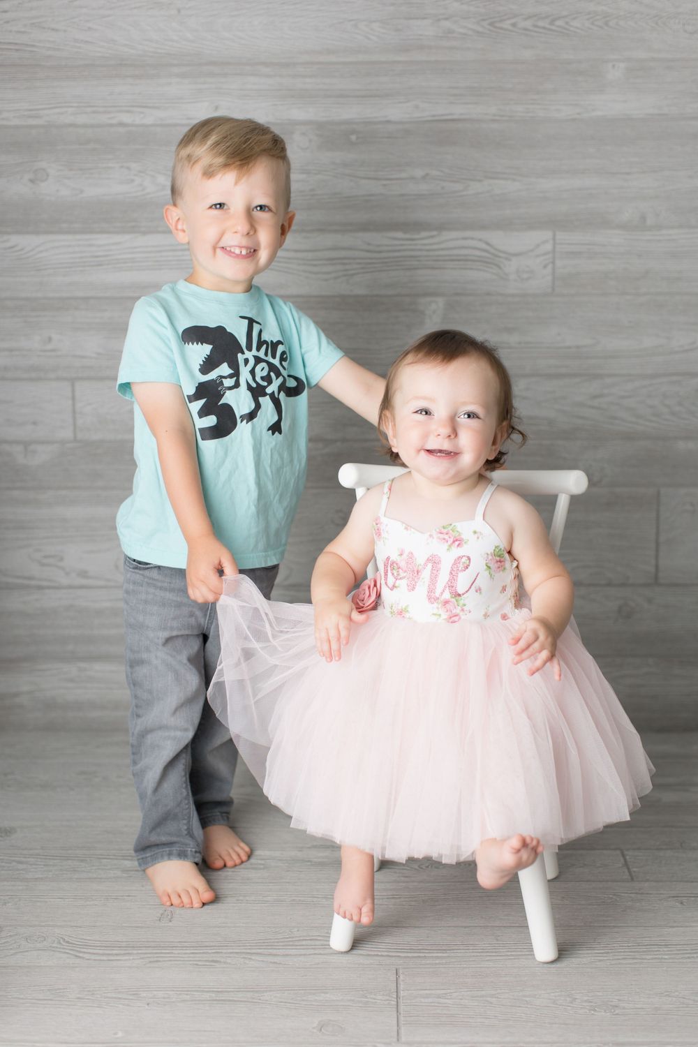 Two children pose together against grey wall, one in tulle dress and one in casual attire.