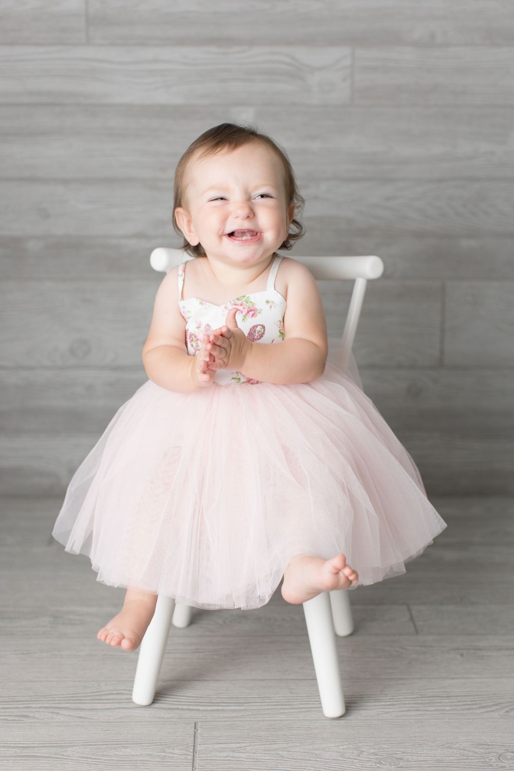 Child in pink tulle dress sits happily on white wooden chair against grey backdrop.