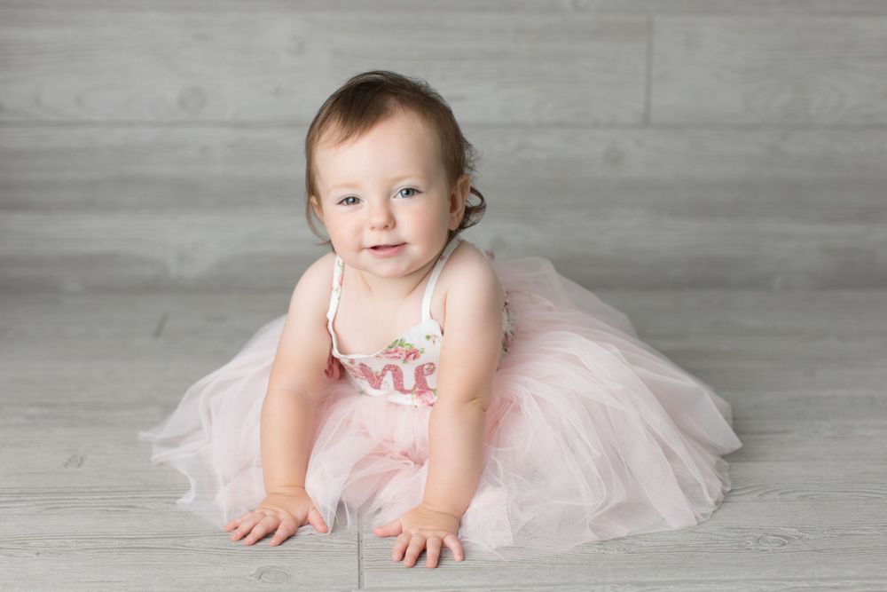 Baby in light pink tulle dress sitting on gray floor during professional photoshoot.