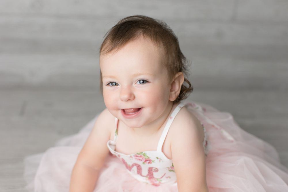 Baby wearing pink tulle dress smiles during portrait session against gray background.