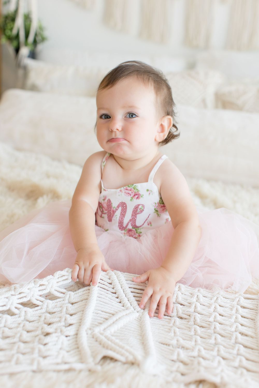 Baby in pink tulle dress sits on textured white blanket during indoor photoshoot.