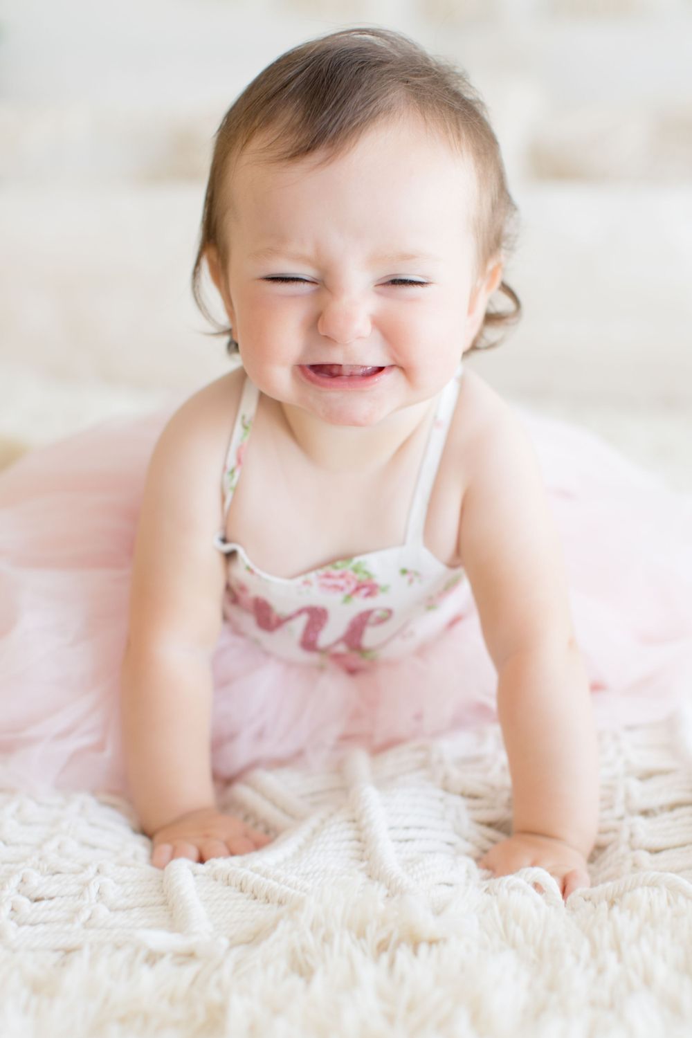 Baby in pink dress laughs with closed eyes while crawling on white blanket.
