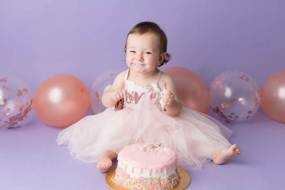 Baby in pink dress sits with birthday cake surrounded by pink and clear balloons.