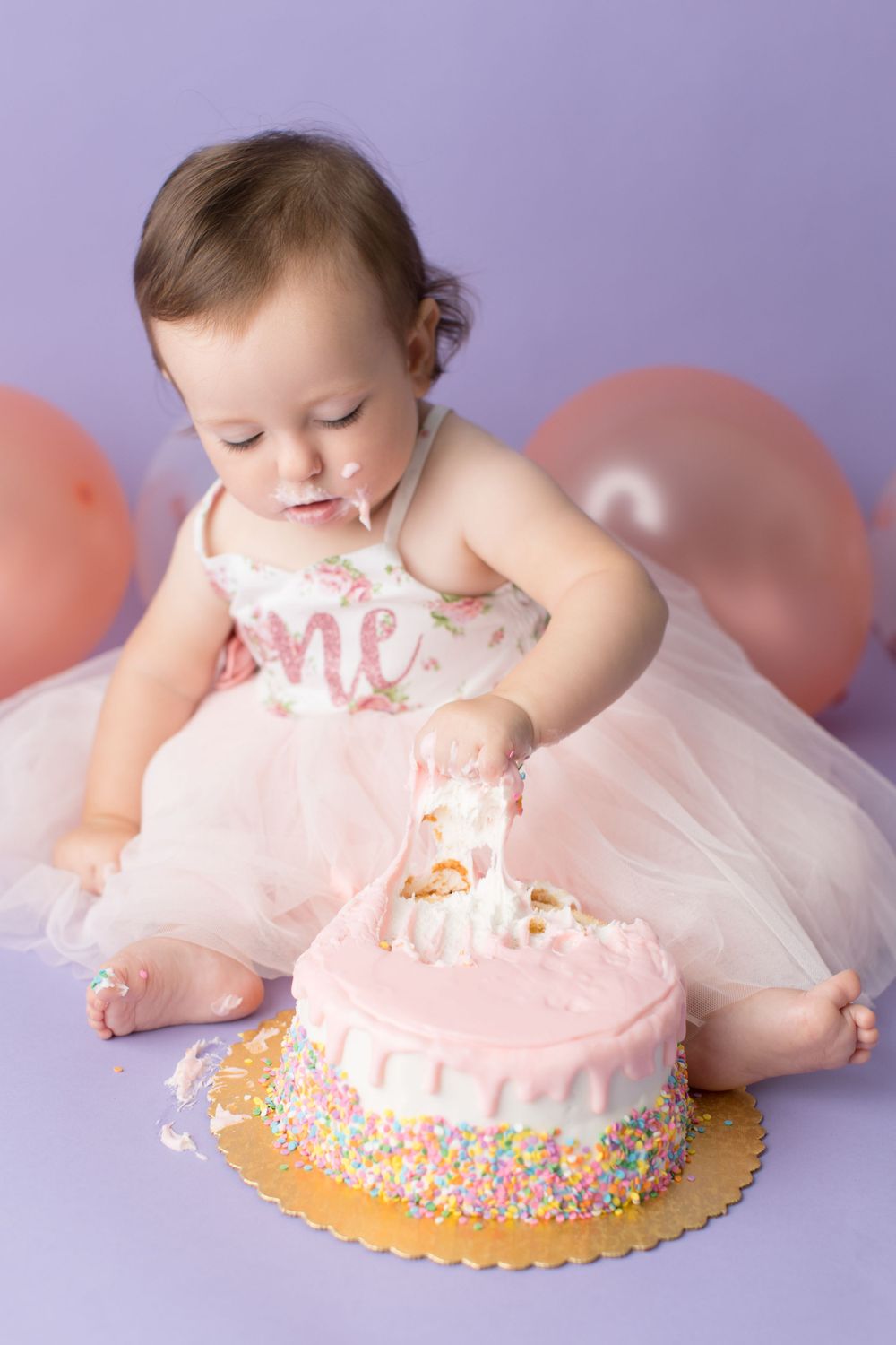 Baby in tulle dress reaches for pink birthday cake during professional photoshoot.