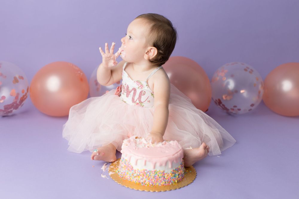 Baby in pink dress explores birthday cake with purple backdrop and balloons.