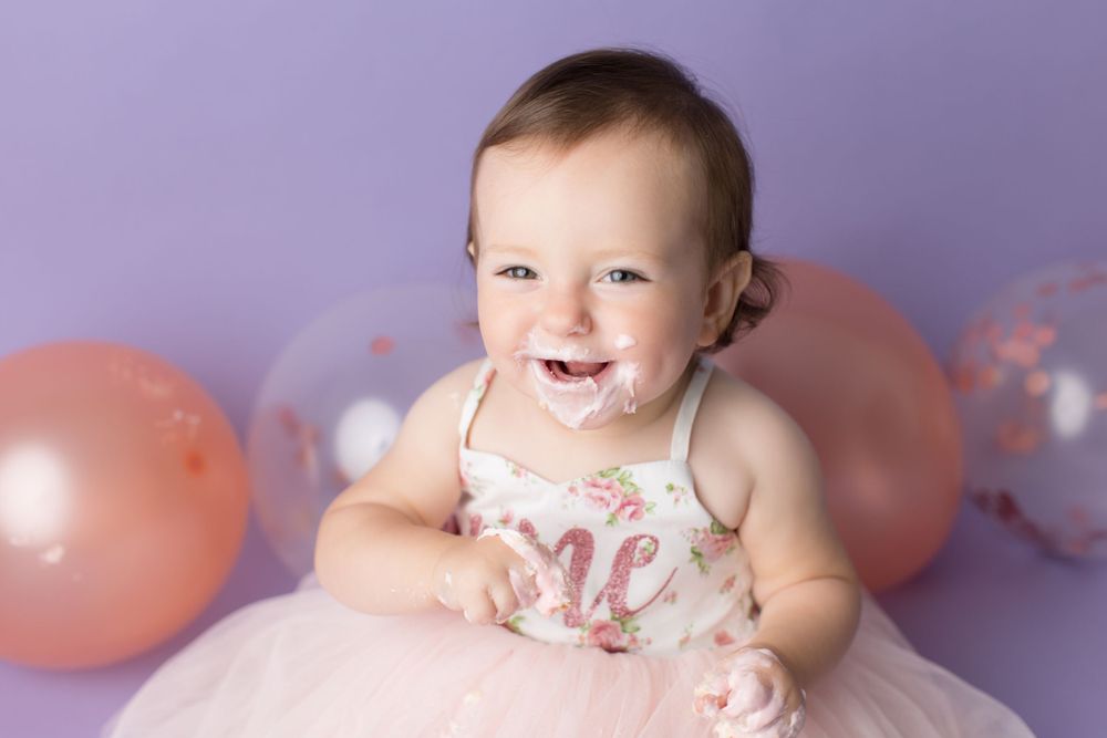 Toddler in pink tutu dress smiling with pink and clear balloons against purple backdrop.