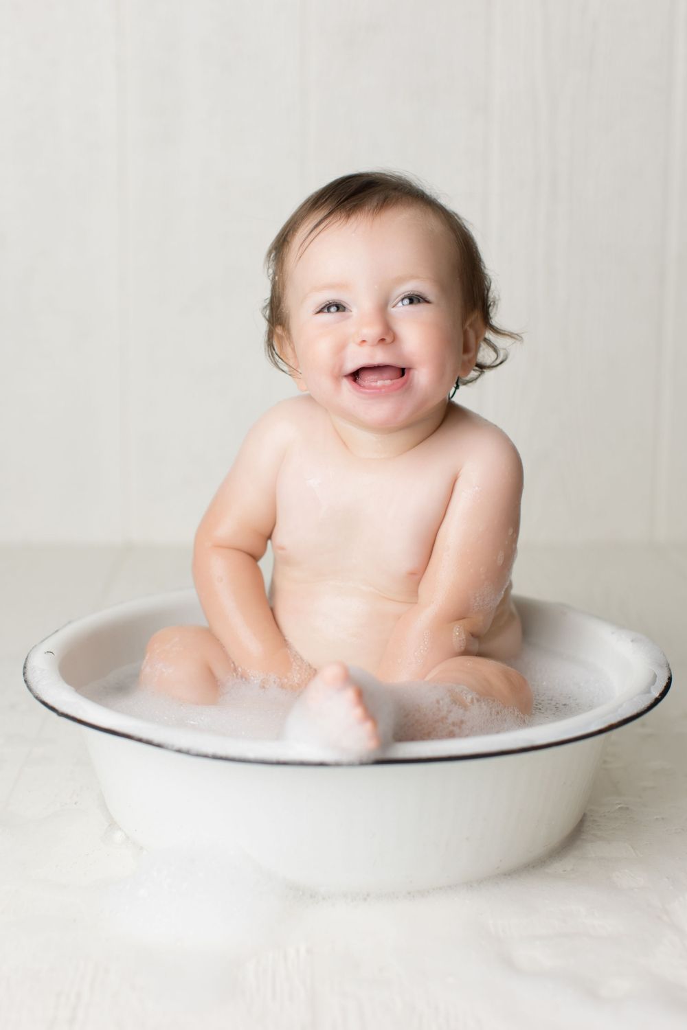 Joyful bath time moment captured in white enamel wash basin against neutral background.
