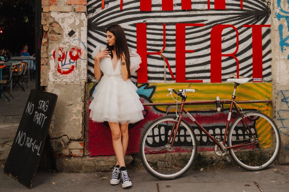 Model in white tulle dress poses against urban graffiti wall with vintage bicycle.