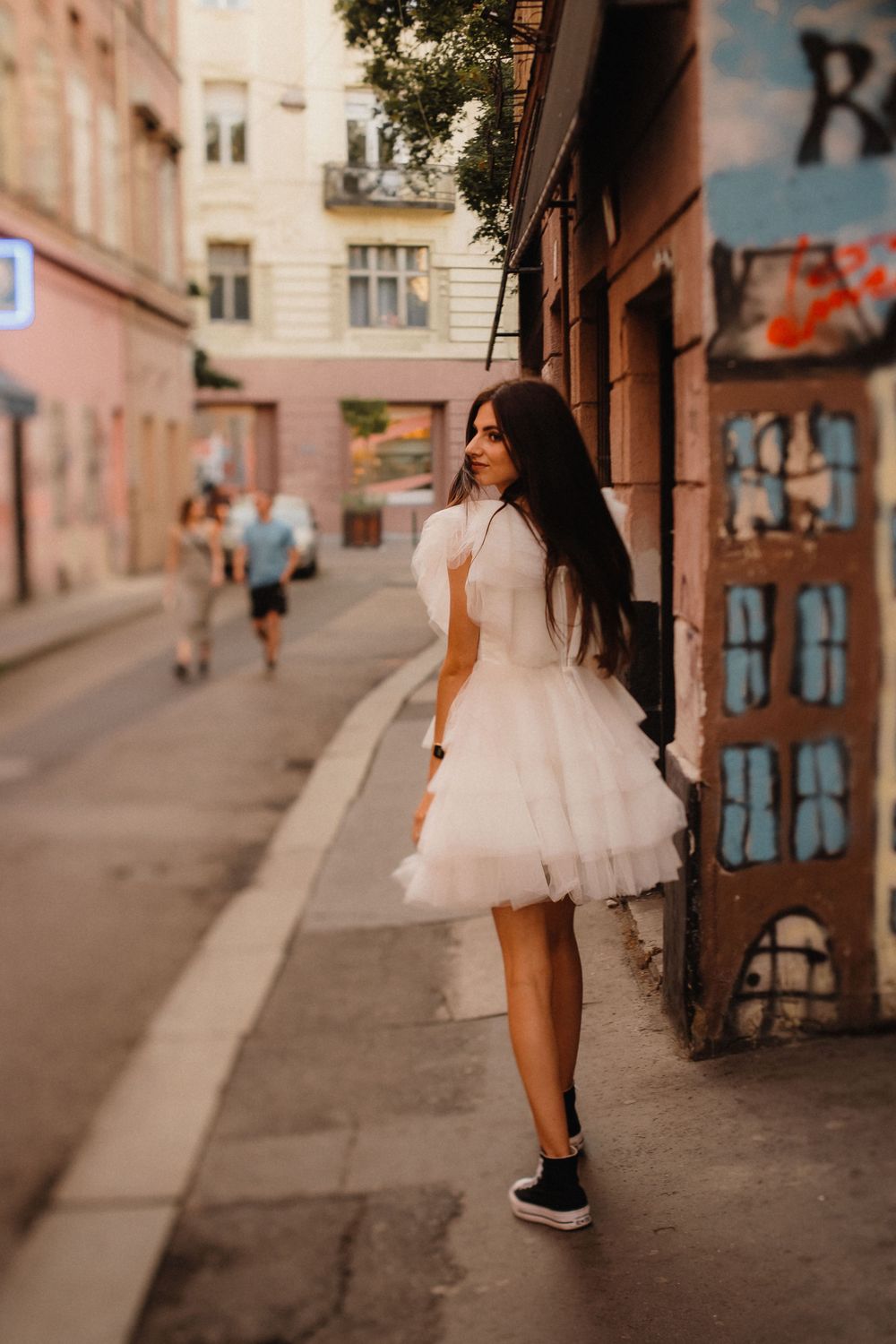 Woman wearing white tulle dress and sneakers poses on city sidewalk near graffiti wall.