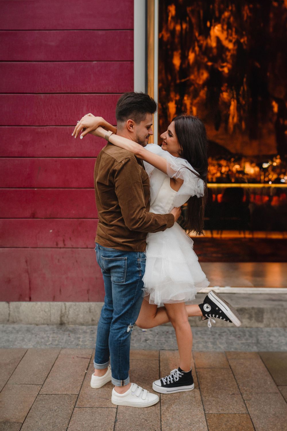A romantic embrace against a red wall with casual sneakers and a white dress creating contrast.