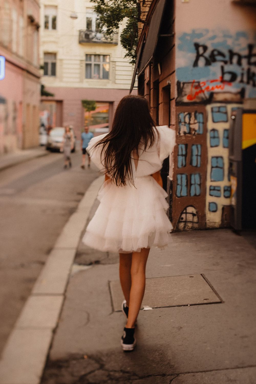 Woman in white tulle dress and sneakers walks down urban street with graffiti wall.