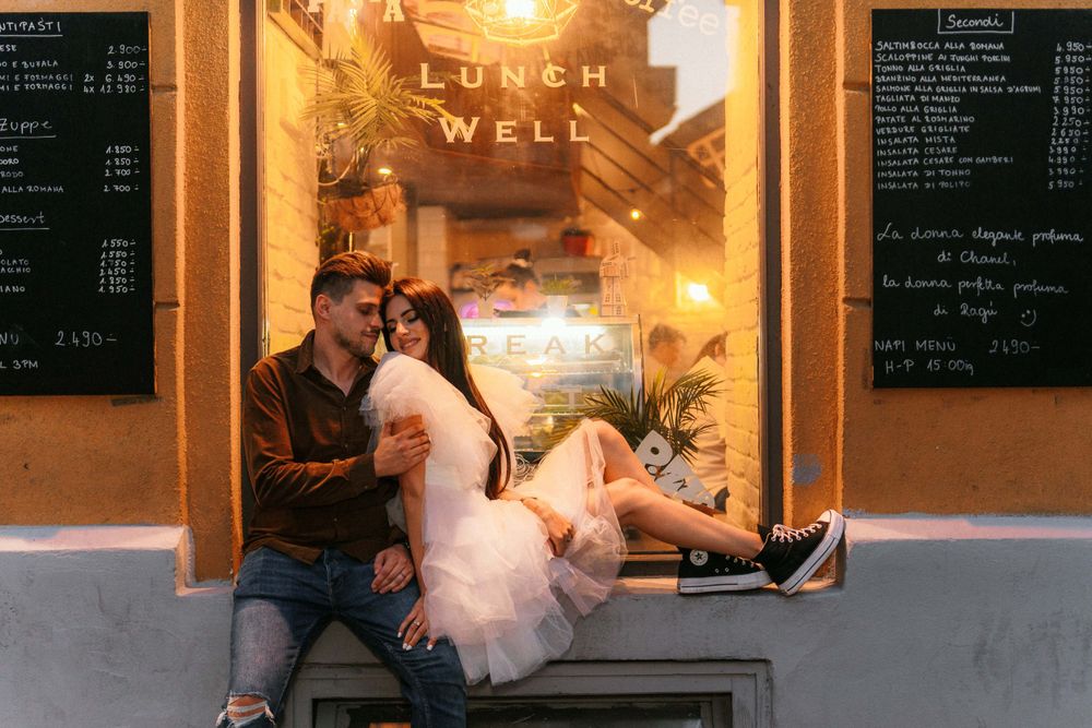 Couple poses on bench outside cafe window with menu boards visible.
