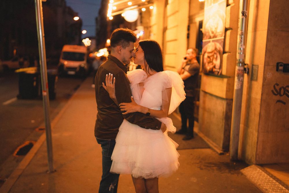 A romantic couple embraces on a dimly lit city street at night beside illuminated storefronts.