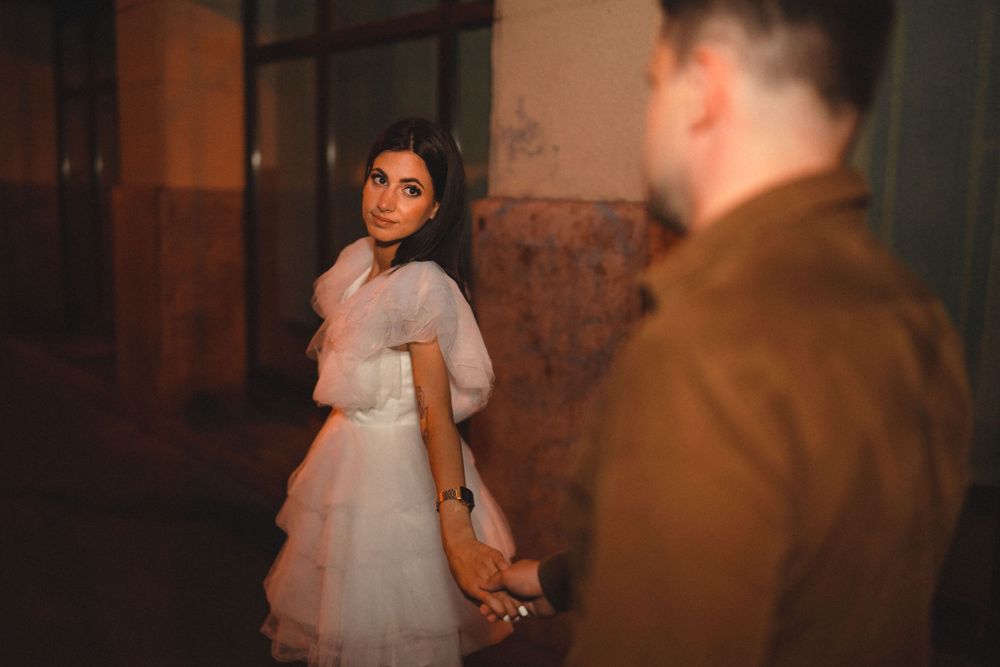 A woman in a white tulle dress glances back while walking on a dark urban street.