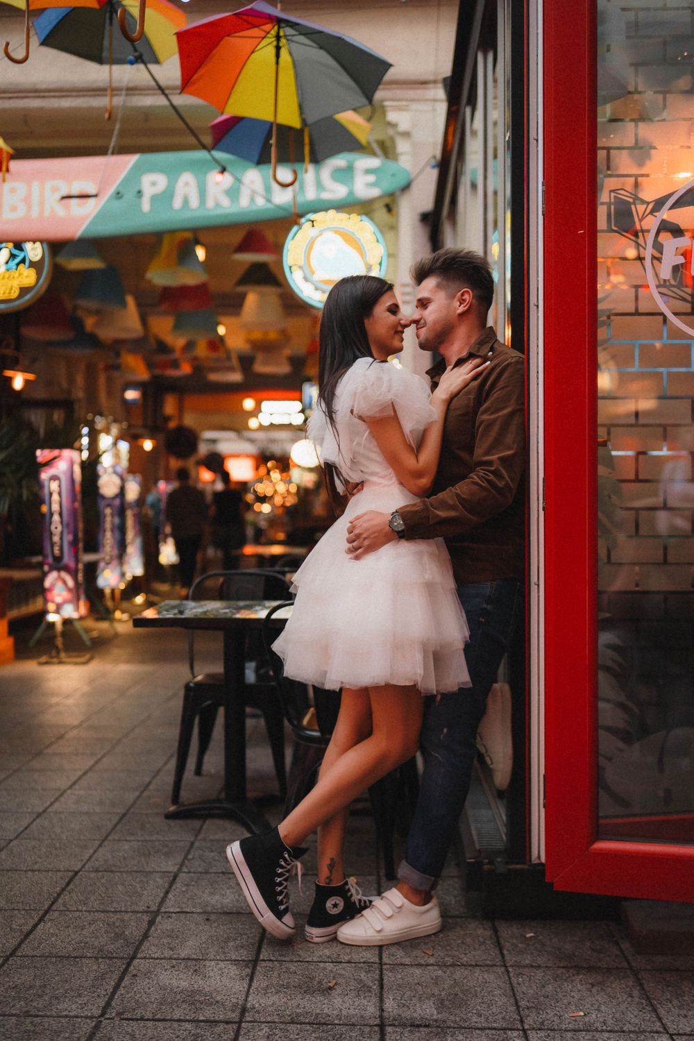 A romantic moment under colorful storefront signs and umbrellas in an urban setting.