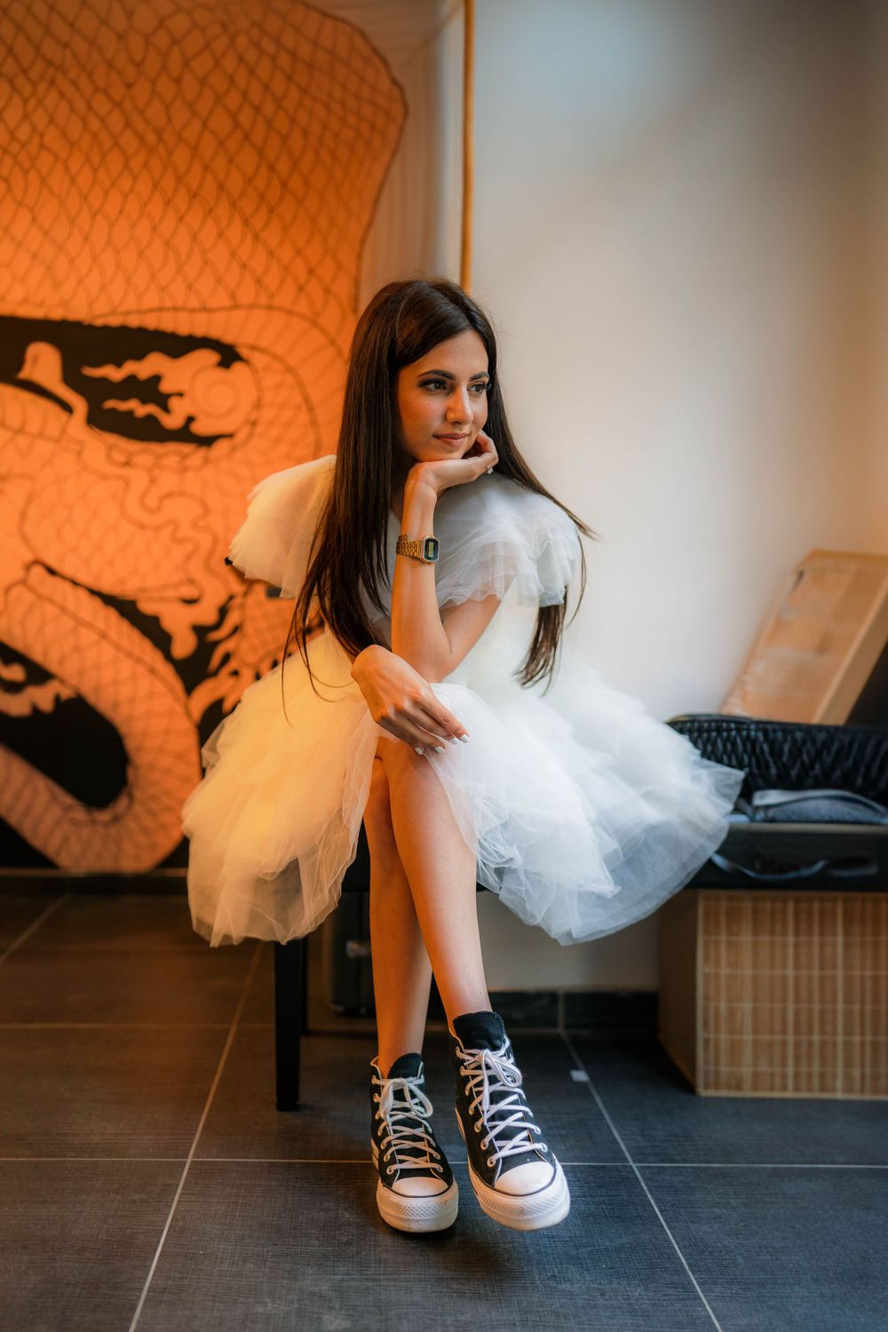 Model in white tulle dress and sneakers poses against orange patterned wall.