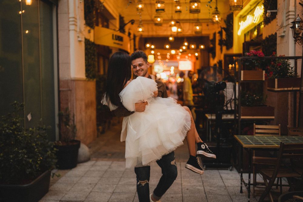 Person carrying someone wearing white tulle dress through illuminated city alleyway.