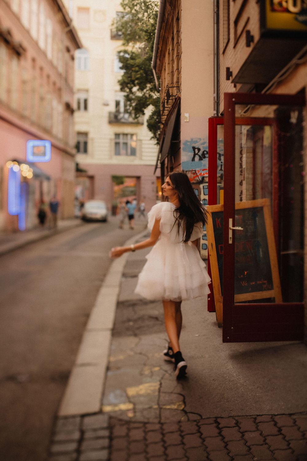 A woman in a white tulle dress walks along a charming European street with storefronts.
