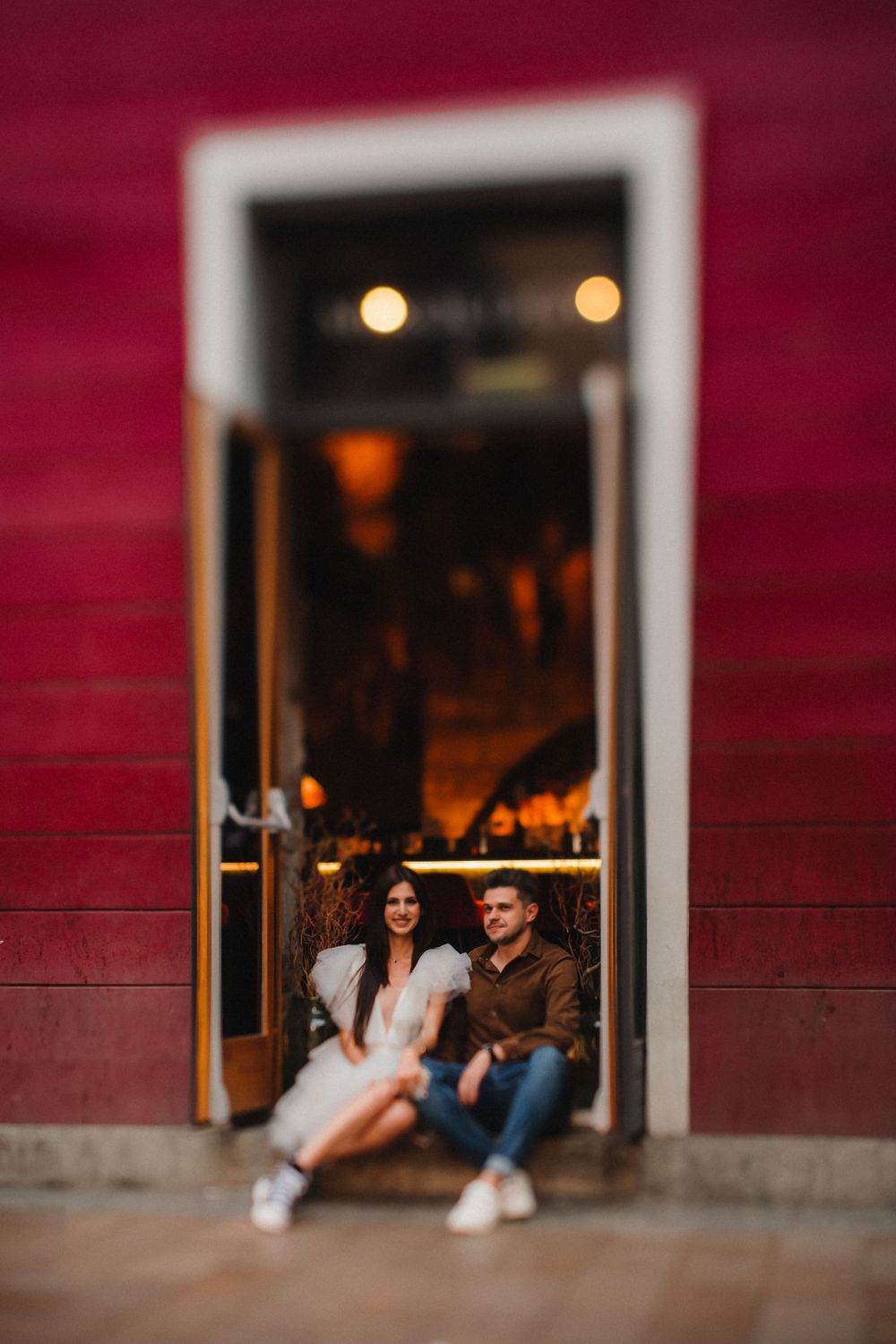 A couple sits in a doorway framed by a red wall during an intimate photoshoot.