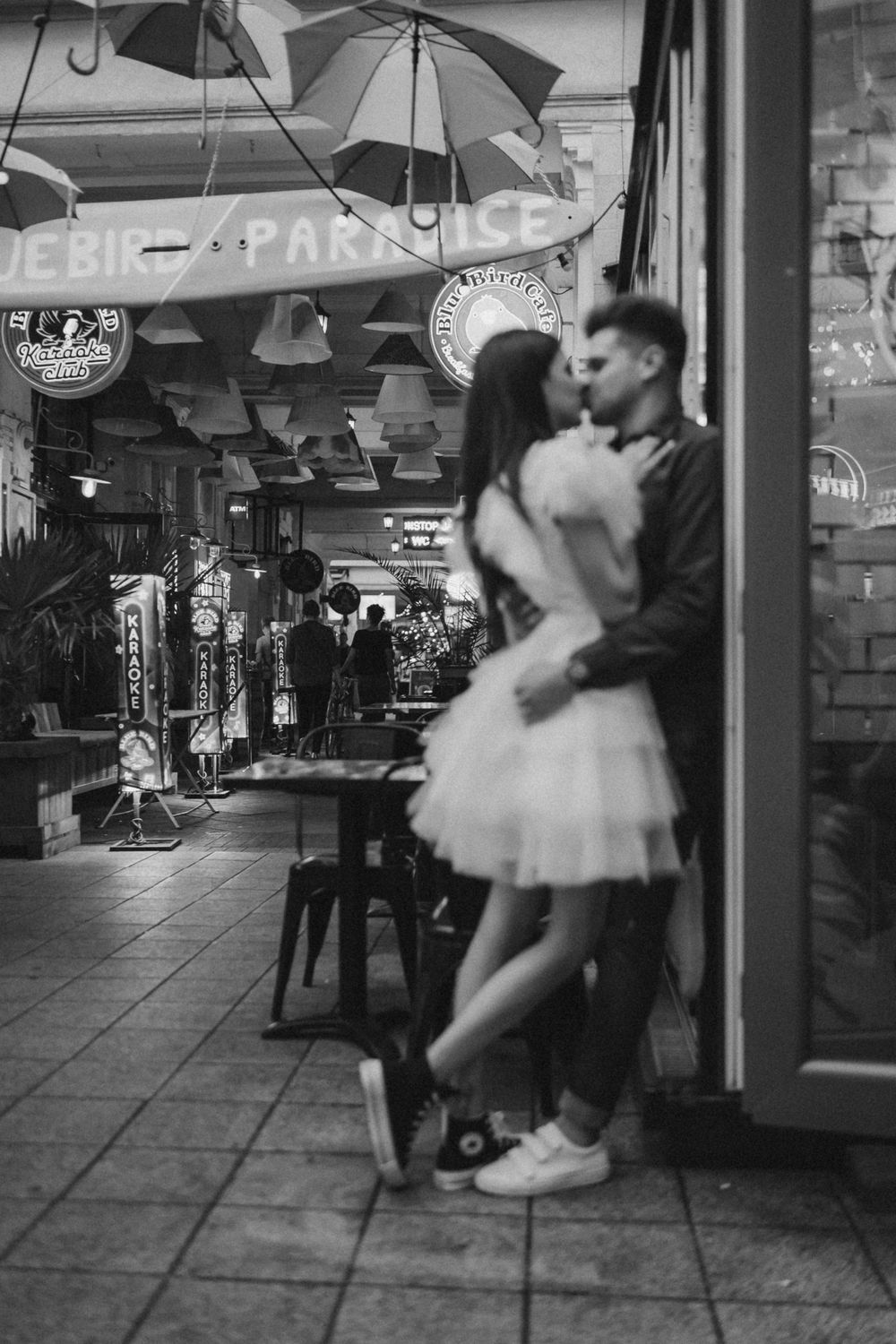 A black and white photo of a couple embracing under hanging umbrellas in a shop corridor.
