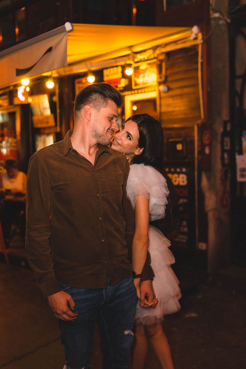 A couple shares a tender moment outside a warmly lit storefront at night.