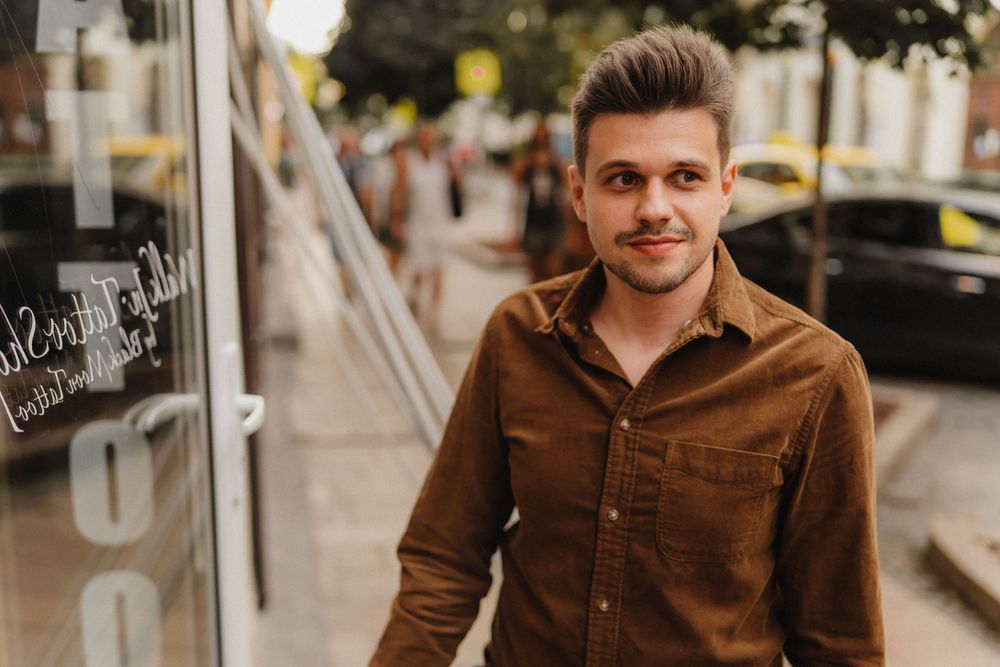 Person in brown shirt standing on a city sidewalk near storefront windows.