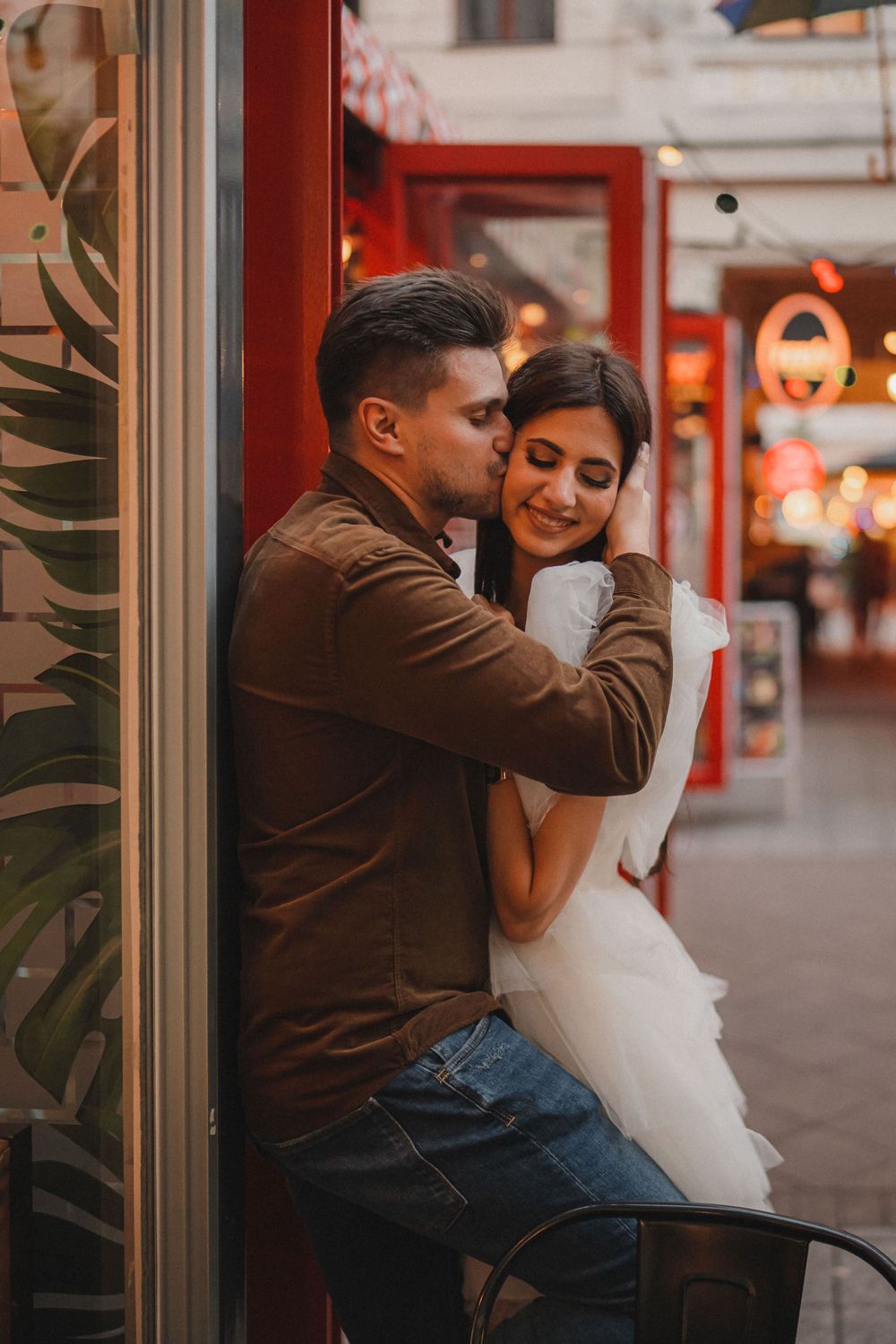 An intimate embrace outside a storefront with warm lighting and tropical plants.