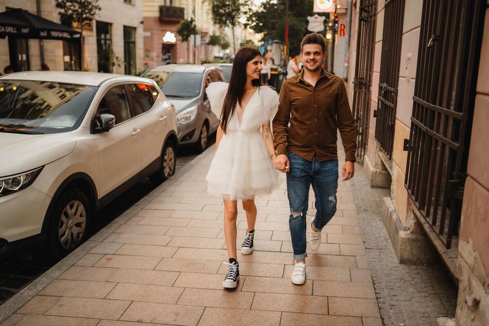 Couple walking together on city sidewalk with parked cars in background.