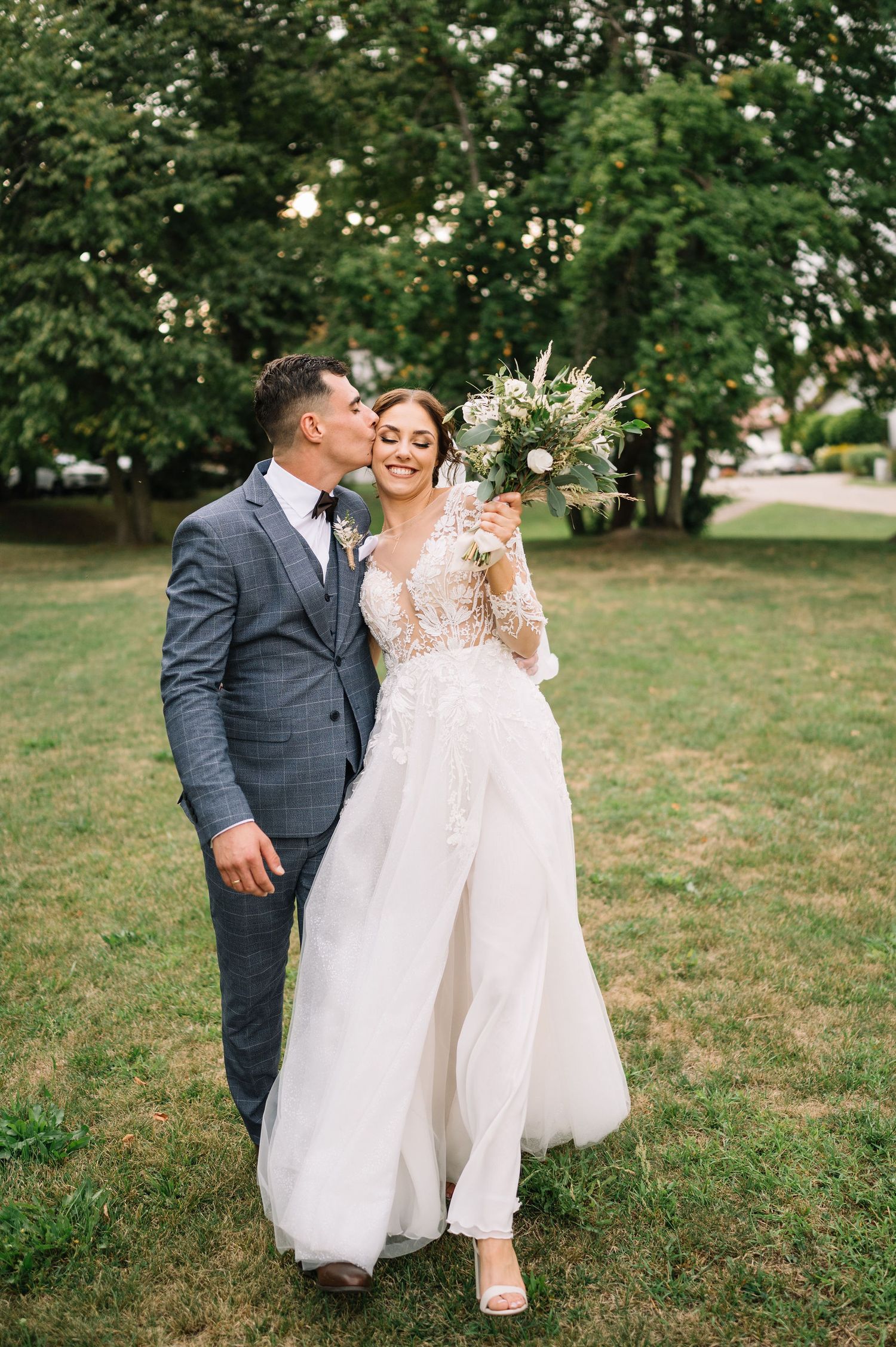 A wedding couple embraces outdoors with the bride holding an elegant white floral bouquet.