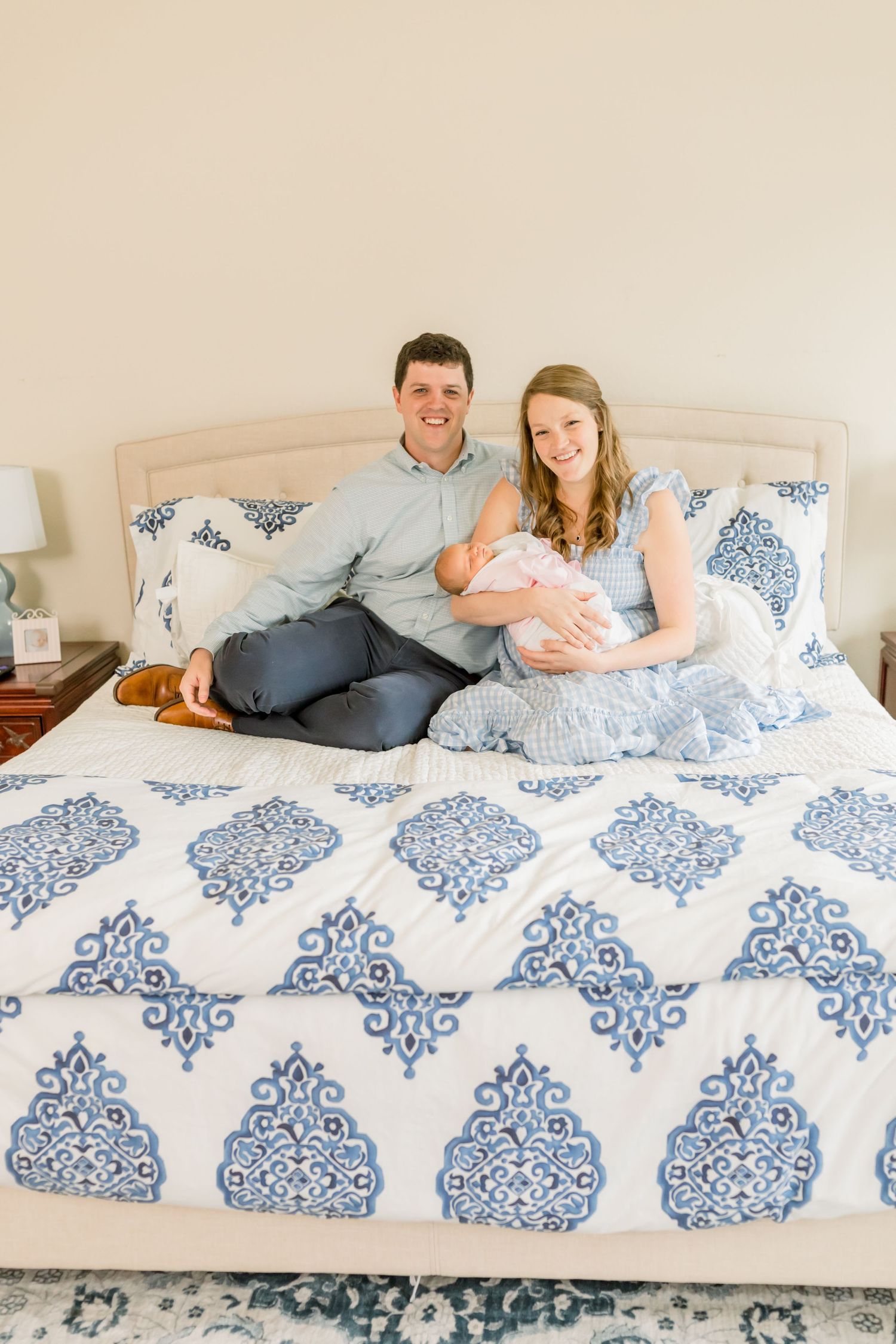 A couple sits together on a bed with blue and white patterned bedding while holding their newborn baby.