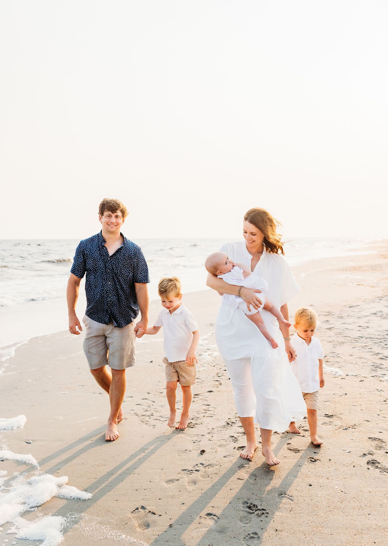 A family walks together along a sandy beach at sunset casting long shadows on the shoreline.