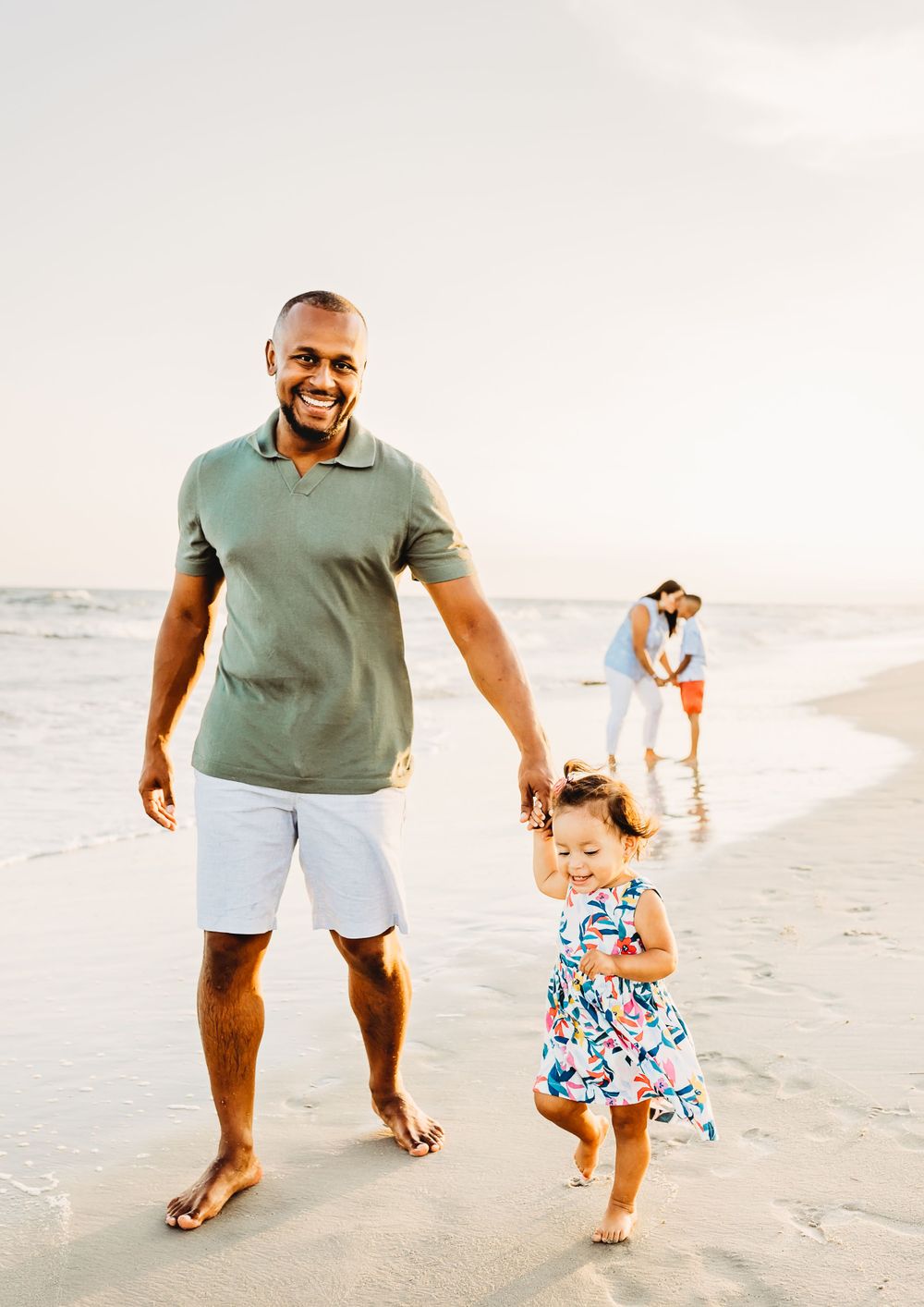 A family enjoys a sunny day walking together on the beach with ocean waves and sand in the background.