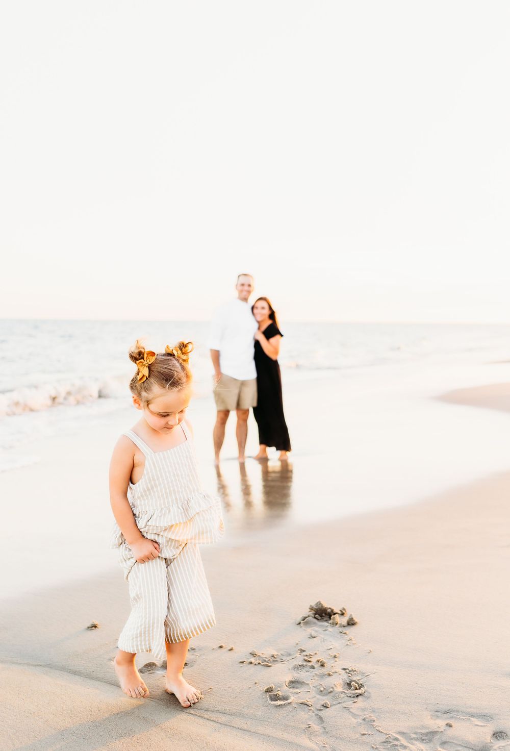 Young child in a light-colored dress plays and explores in the sand along a peaceful beach shoreline.