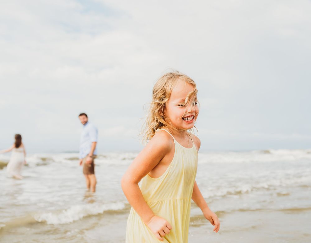 A young child in a yellow dress plays joyfully at the beach on a cloudy summer day.