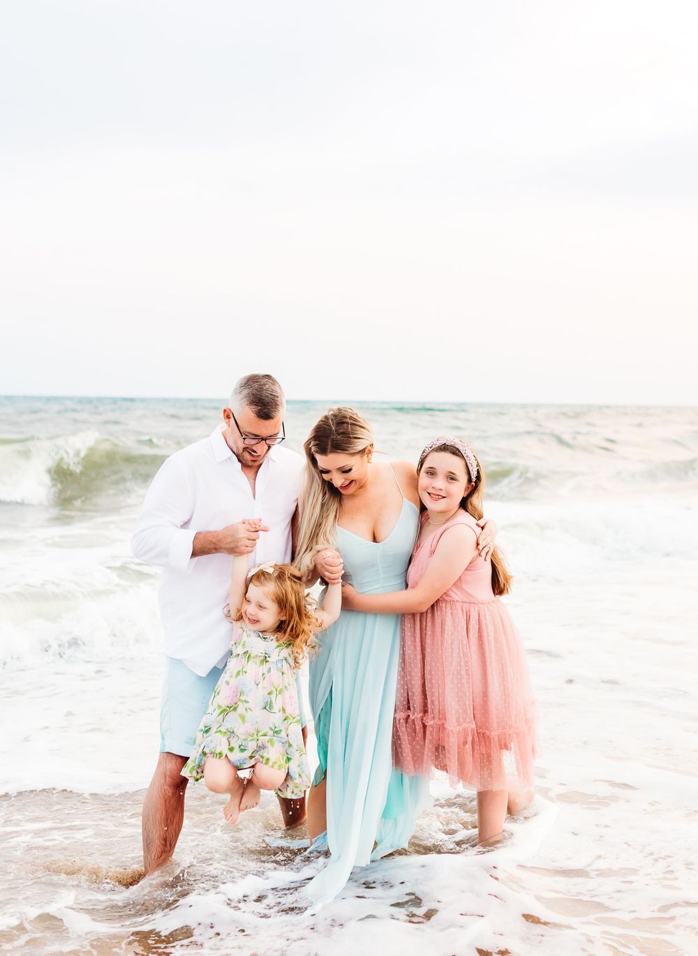 Family enjoying a playful moment together splashing in the ocean waves at sunset wearing pastel dresses and white clothing.