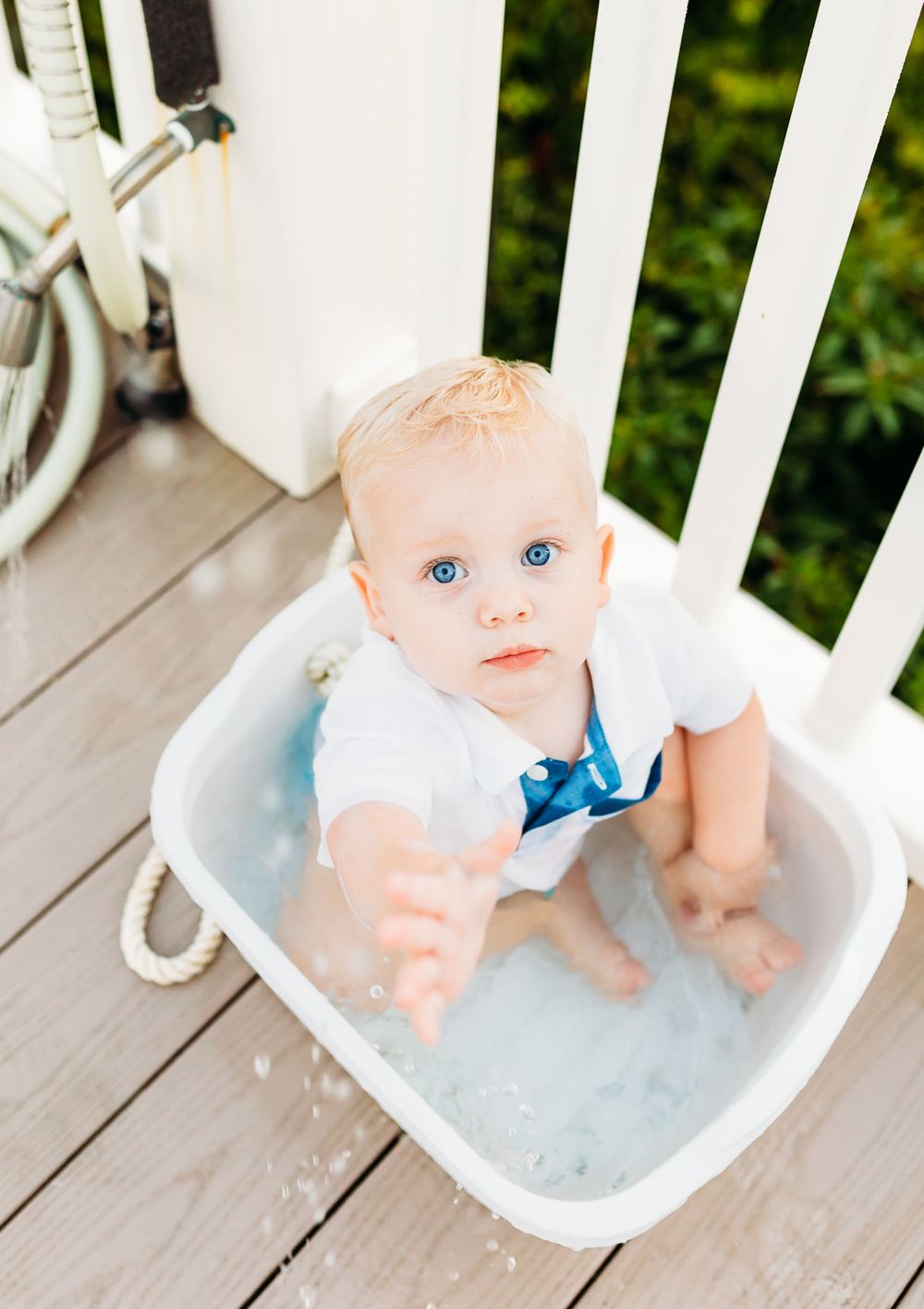 Small baby wearing white outfit with blue bow tie sits in white plastic tub on wooden deck.
