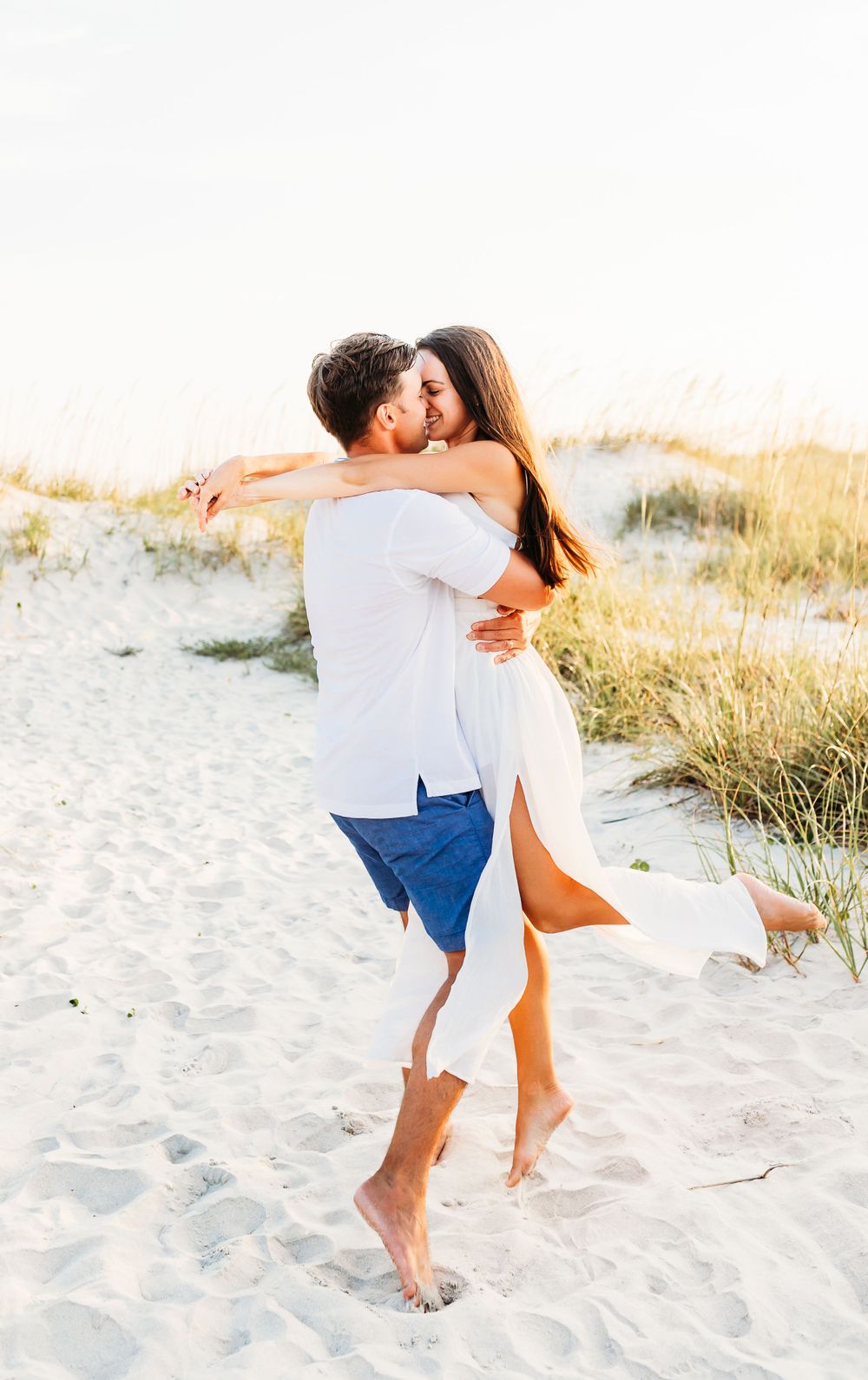 A couple in white outfits embracing and dancing playfully on a sandy beach at sunset.