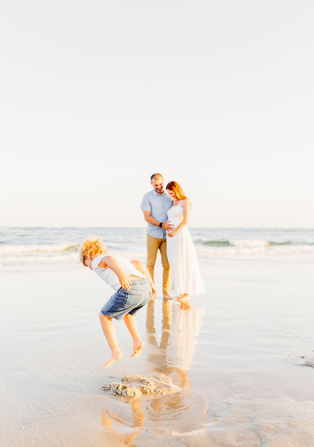 A family enjoys a sunny day at the beach while a child playfully jumps in the shallow ocean water.