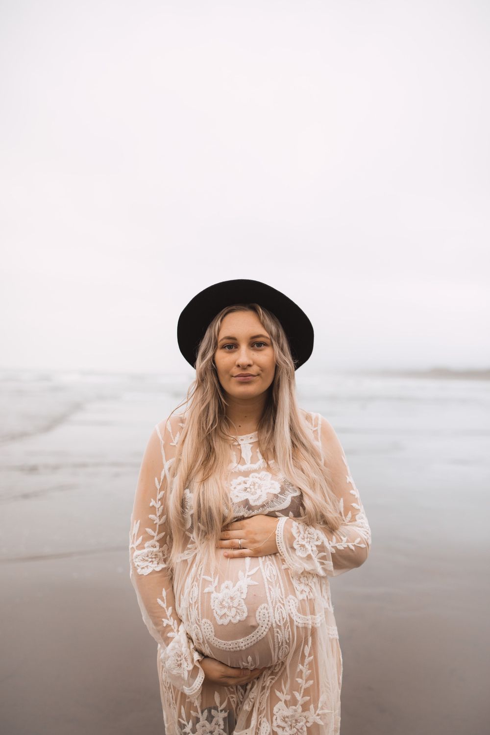 A maternity photo on a misty beach featuring a white lace dress and black hat against a moody gray backdrop.