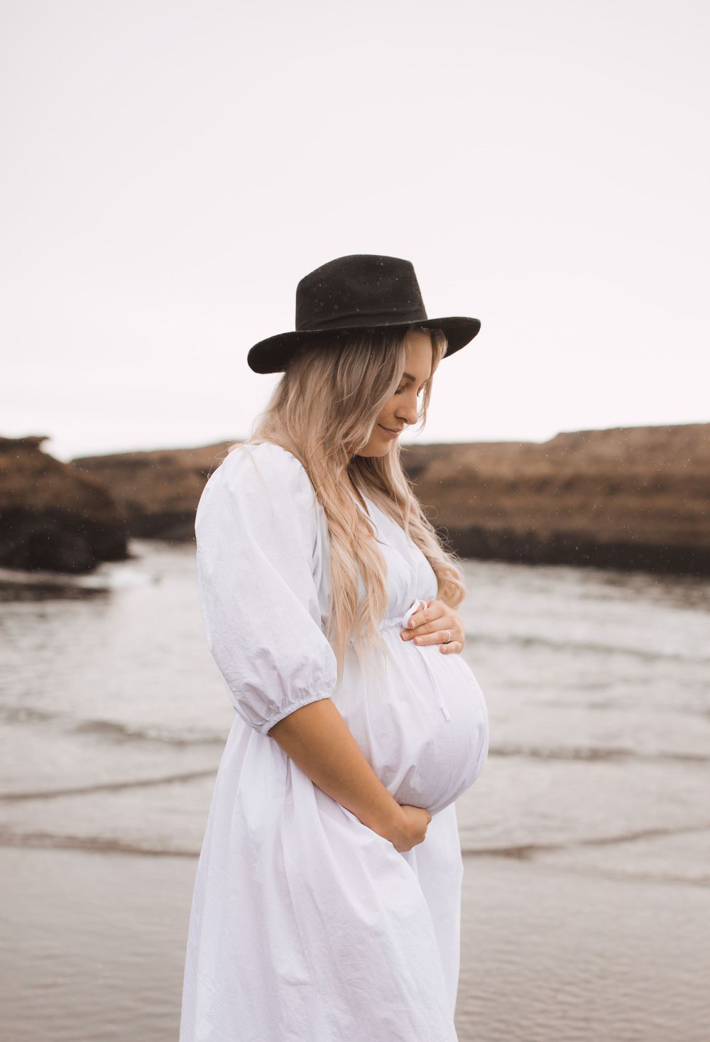 A person in a white dress and hat stands on a rocky beach during a maternity photoshoot.