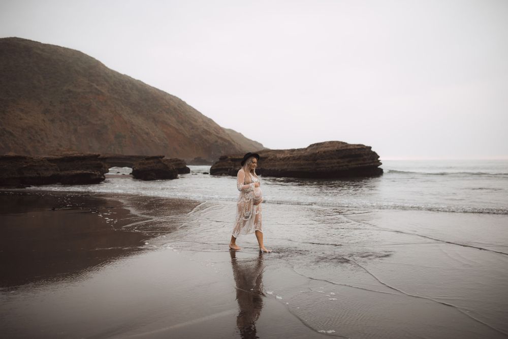 A dreamy beach maternity session captured in soft, muted tones with coastal rocks in the background.