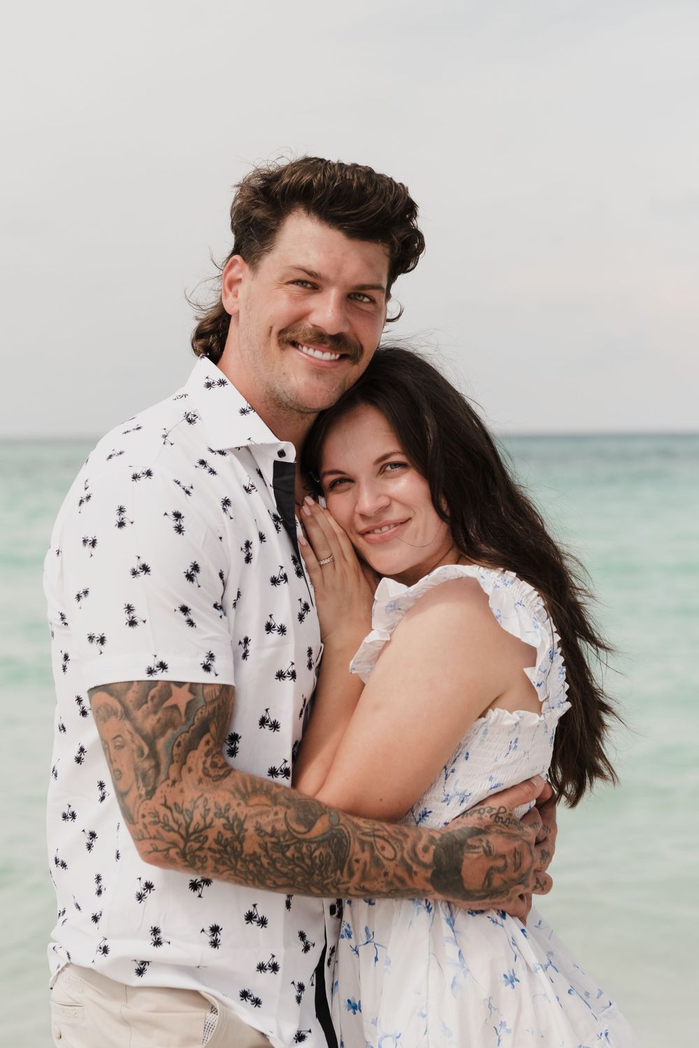 Couple in white summer outfits sharing intimate moment on beach with turquoise water background.