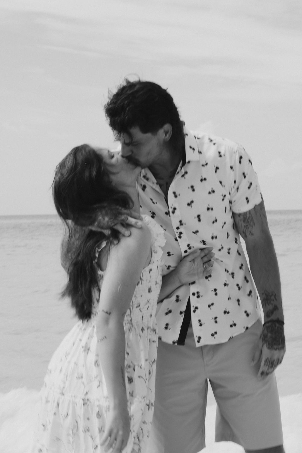 A romantic black and white beach photo of a couple in matching patterned outfits sharing an intimate moment.