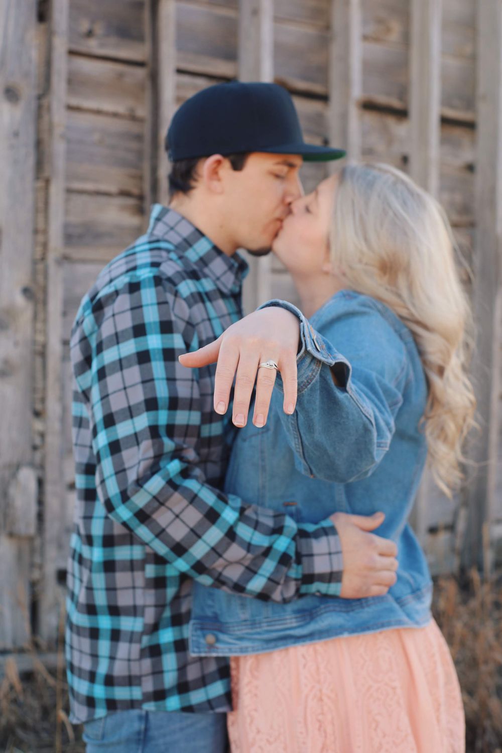 Romantic couple in casual attire sharing a kiss against a rustic wooden backdrop.